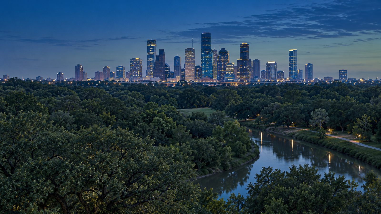 A downtown skyline at dusk beyond a canopy of oak trees and a winding bayou