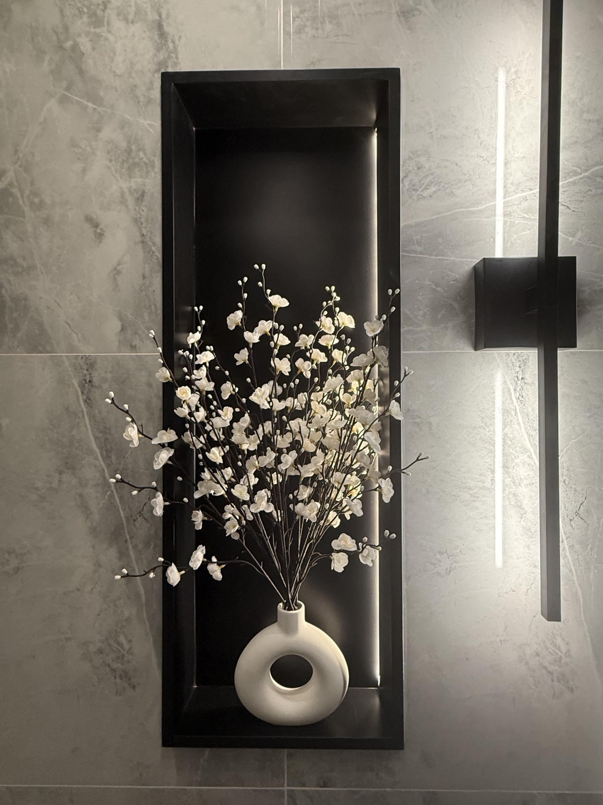 Lit black niche set into grey marble, holding white blossom branches in a round white vase