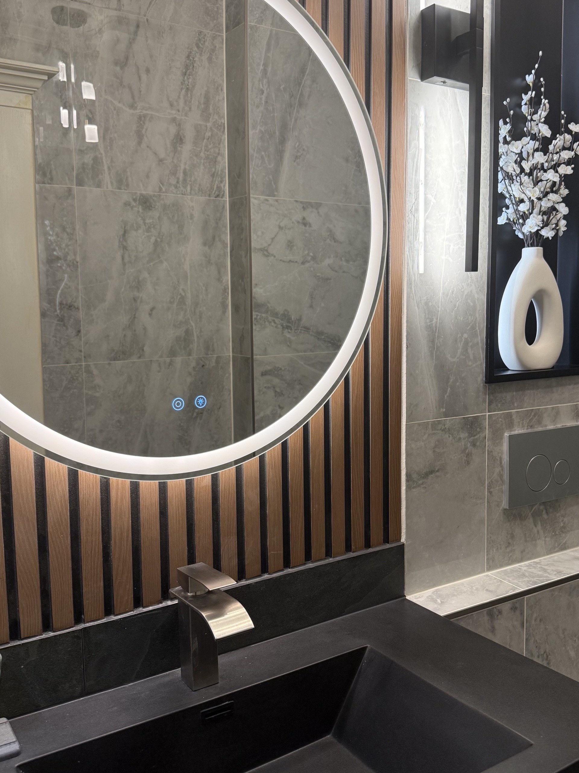 Round backlit mirror against oak slats above a black basin with a brushed nickel faucet